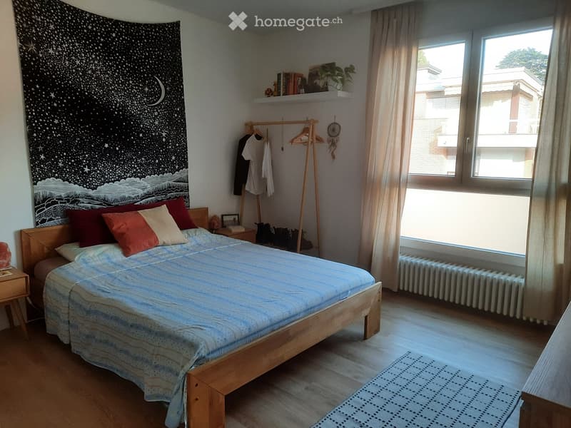 Cozy bedroom with a wooden bed, colorful pillows, a nightstand, a radiator, and a large window with curtains. A wooden hanger with clothes, a shelf with books and a plant, and a dreamcatcher are also visible.