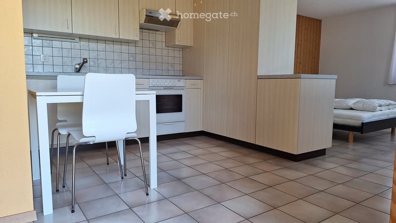 A modern, compact kitchen and living area with white cabinets, a tiled backsplash, a small dining table with chairs, and a bed in the corner.