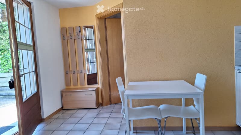 A small, furnished room with a white table and two white chairs, a mirror, and a wooden cabinet with drawers.