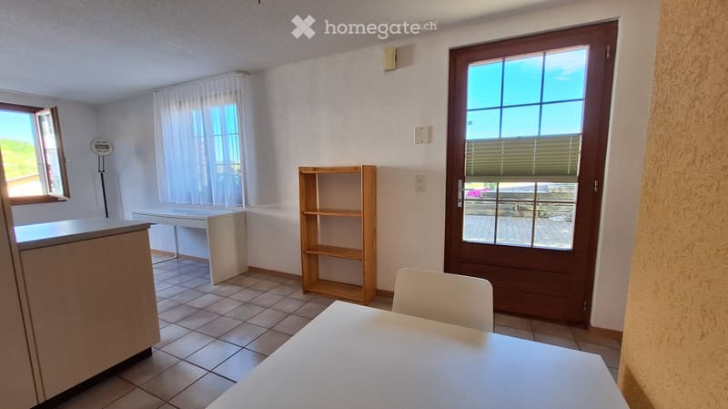 Bright, unfurnished room with tiled floor, large windows, a wooden shelf, a white desk, and a small table with a chair.
