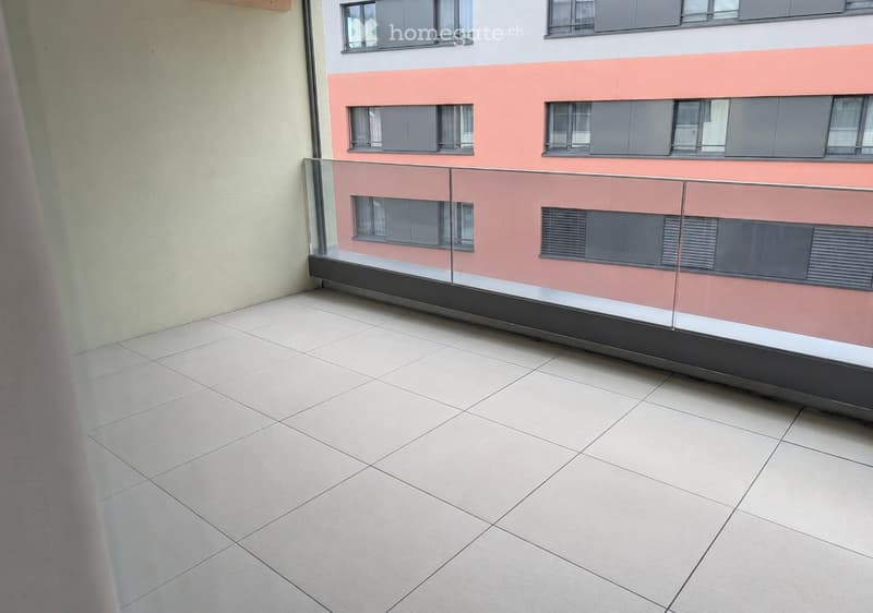 Balcony with tiled floor and glass railing, overlooking a modern building with pink and white facade and multiple windows.