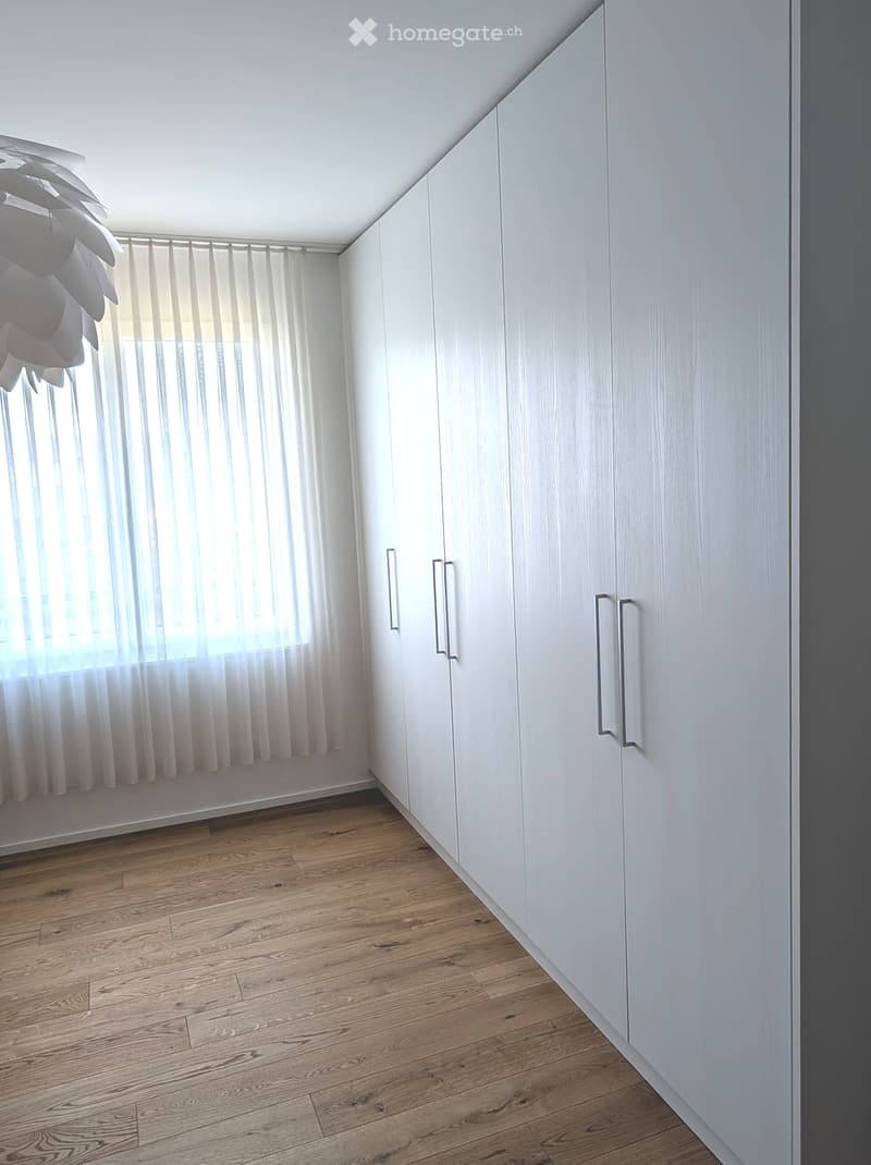 Empty room with light wooden flooring, white built-in wardrobes, and a window with sheer curtains.