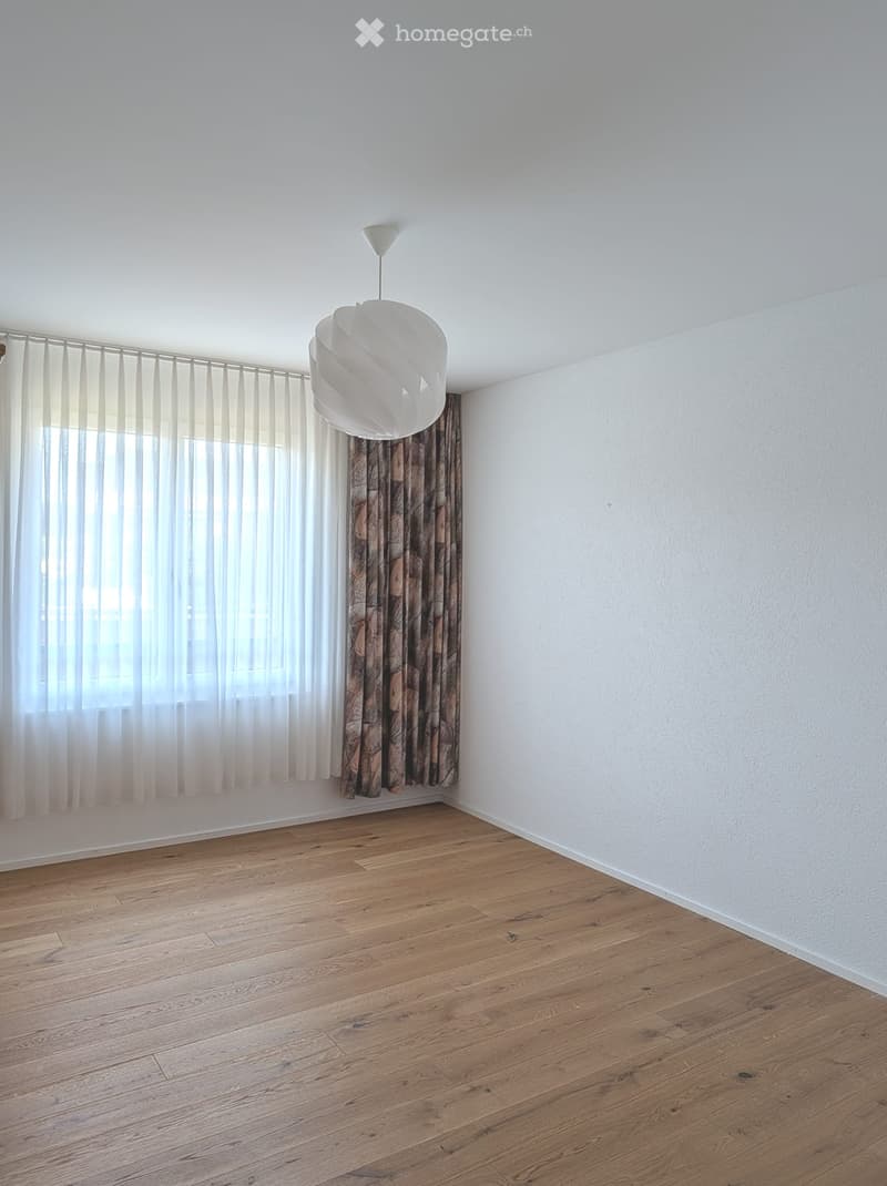 Empty room with wooden flooring, white walls, and a large window with sheer curtains and patterned drapes. A modern pendant light hangs from the ceiling.