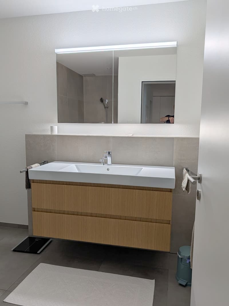 Modern bathroom with a double sink, wooden cabinet, large mirror with integrated lighting, towel, and trash bin.