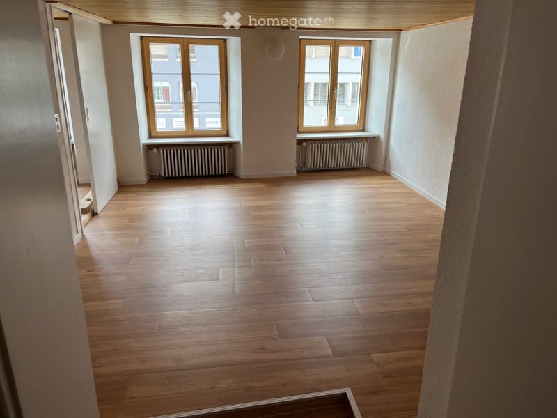 Empty room with wooden flooring, large windows, radiators, and a visible doorway leading to another space.