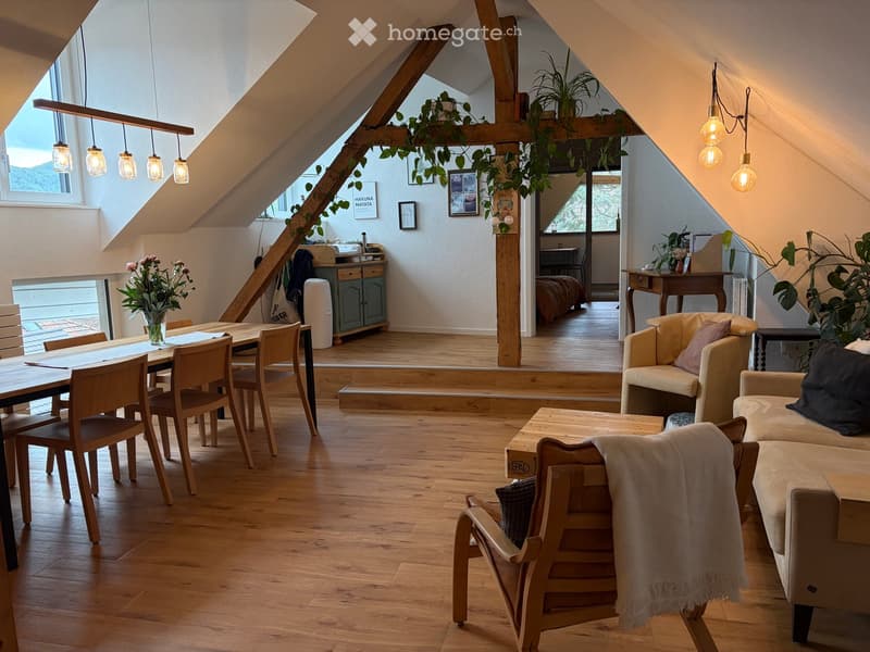 Bright, open living space with wooden floors, exposed wooden beams, and large windows. Features a sofa, coffee table, dining table with chairs, and decorative plants.