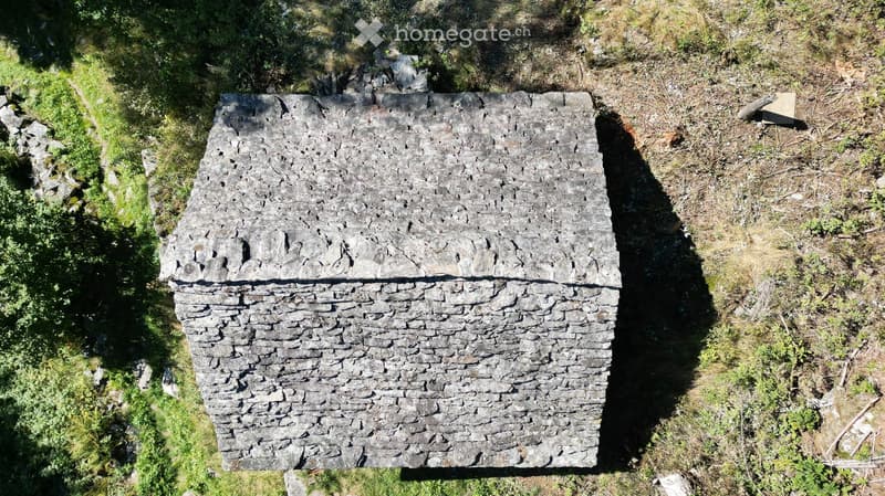 An aerial view of a stone structure, likely a small building or shed, situated on a grassy, elevated terrain surrounded by vegetation and scattered rocks.