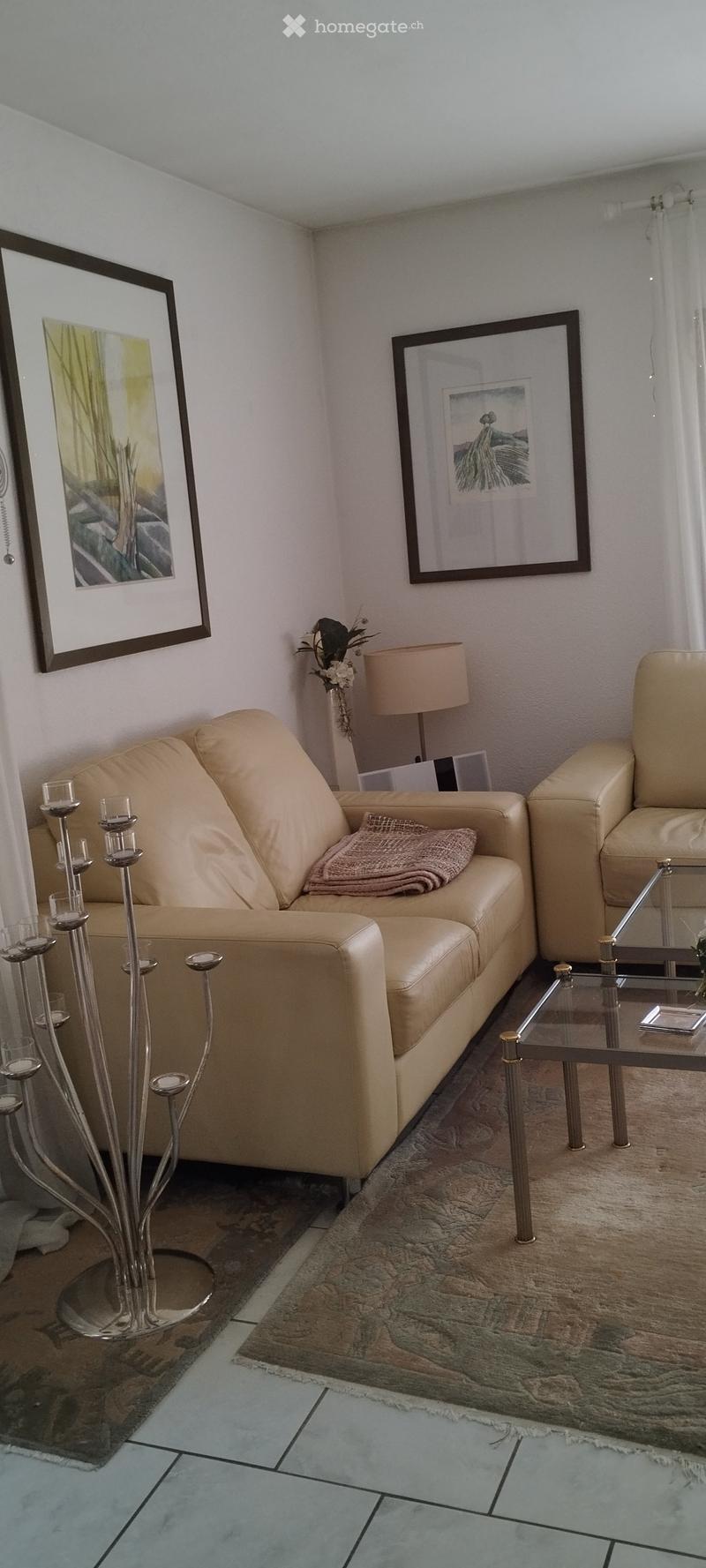 Living room with a beige leather sofa, matching armchair, glass coffee table, table lamp, candle holder, and two picture frames on the wall.