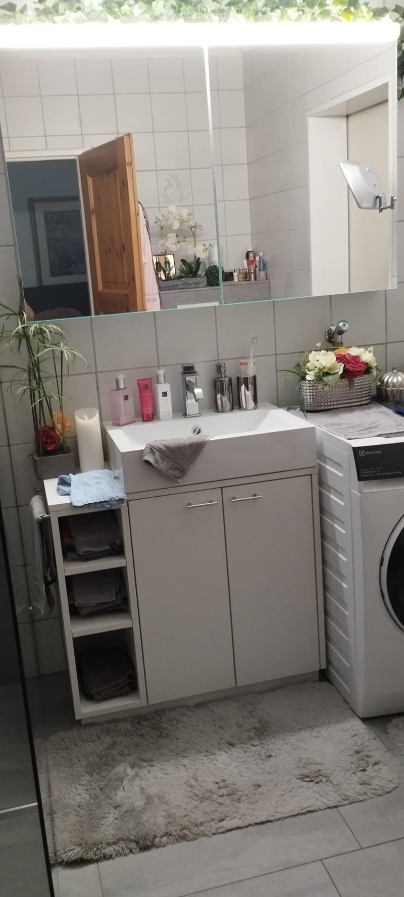 Modern bathroom with a white sink, mirror, cabinet, and washing machine. Includes a towel rack, flower arrangement, candle, and various toiletries.