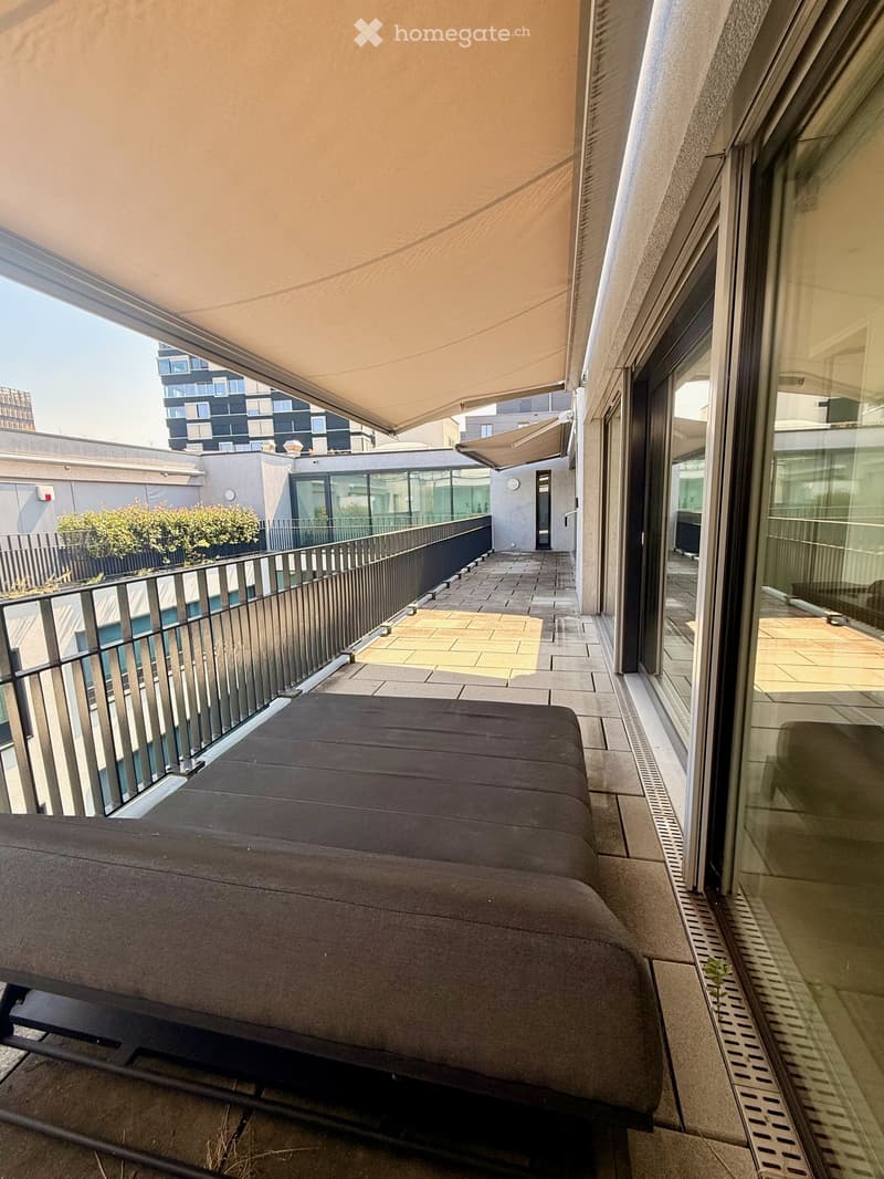 Balcony with a grey sofa, modern railing, and a retractable awning, overlooking a cityscape with modern buildings.
