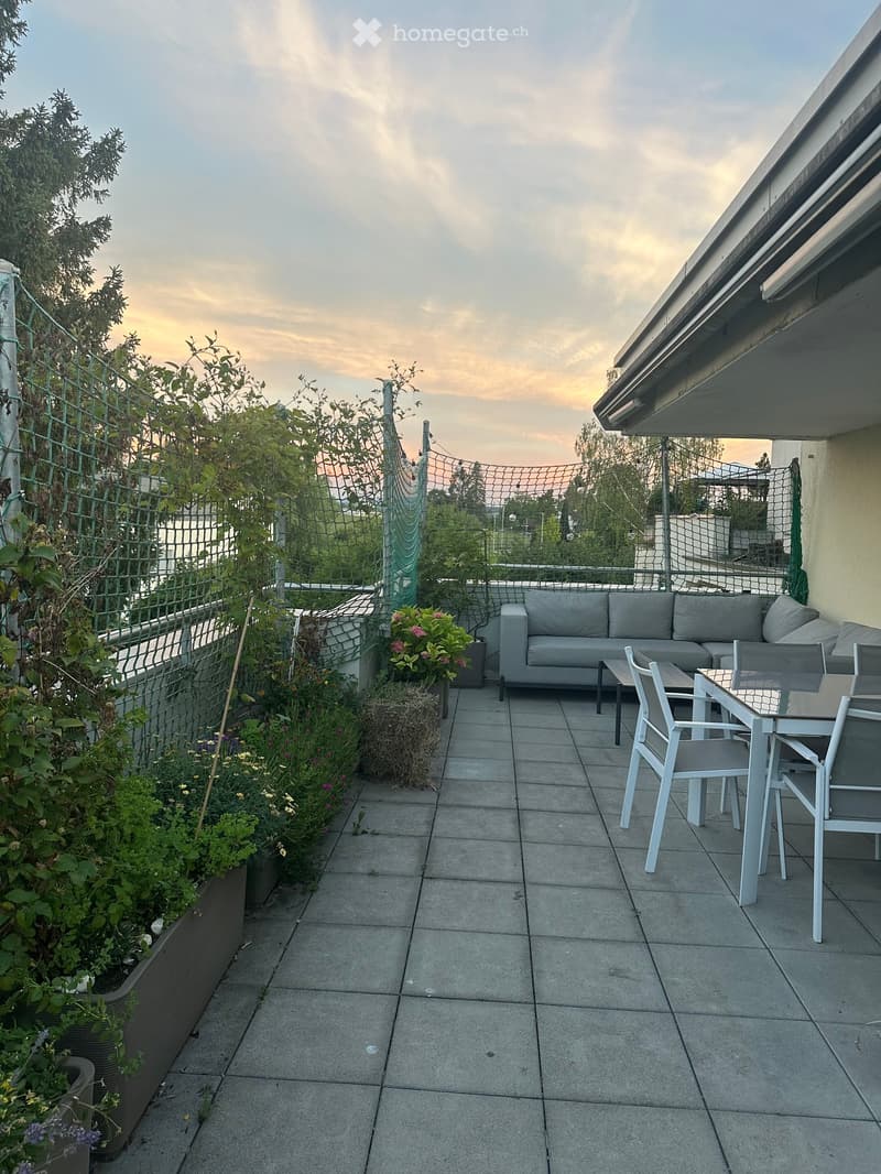Balcony with tiled floor, grey sofa, small table, and chairs, surrounded by greenery and a view of the city.