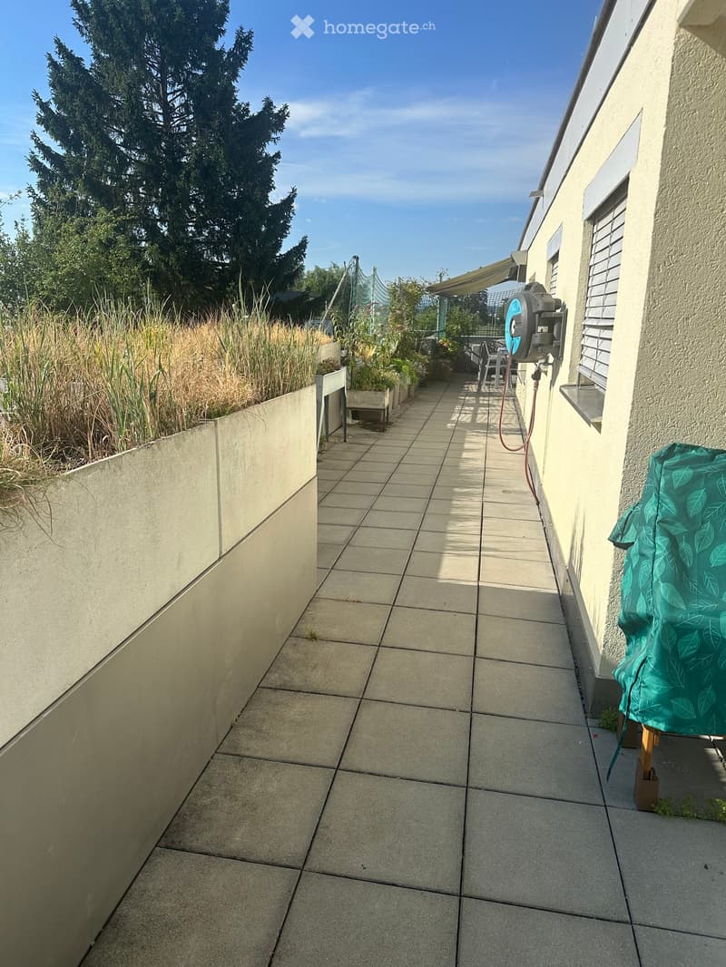 A narrow, tiled outdoor pathway bordered by a low concrete wall on one side and a building on the other, with greenery and a covered seating area visible in the distance.