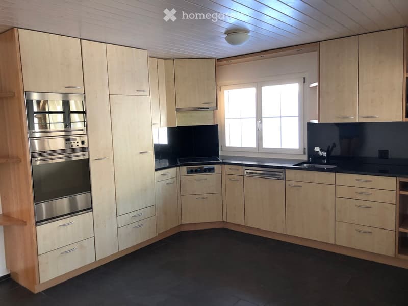 Modern kitchen with light wood cabinets, stainless steel oven and dishwasher, black countertop, and a window above the sink.