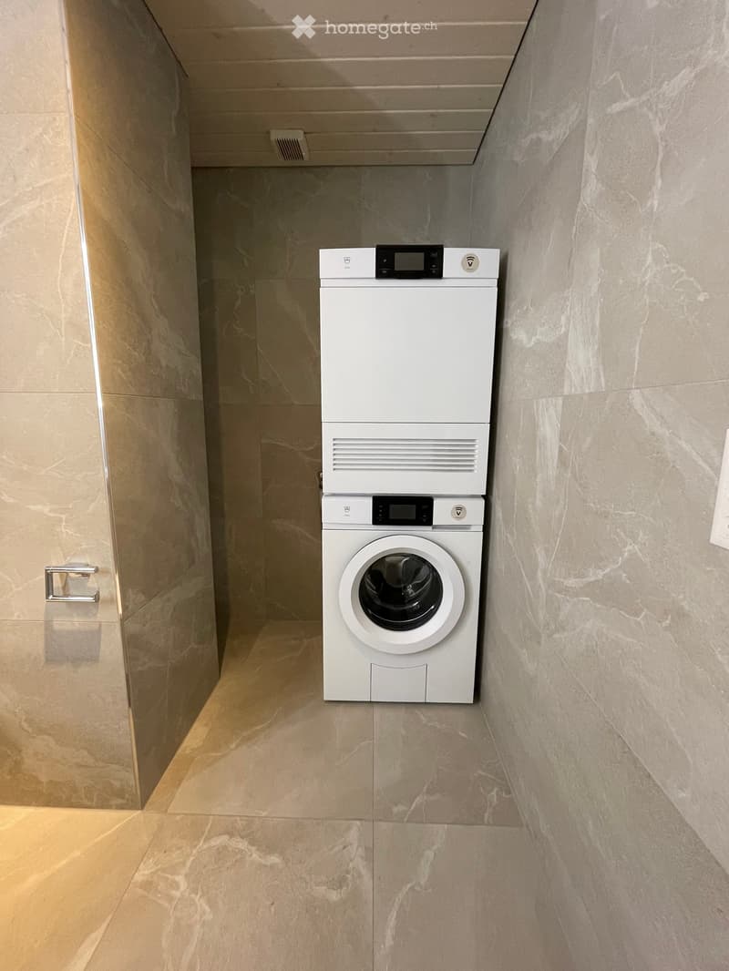 A narrow hallway with marble flooring and walls, featuring a stacked washing machine and dryer unit.