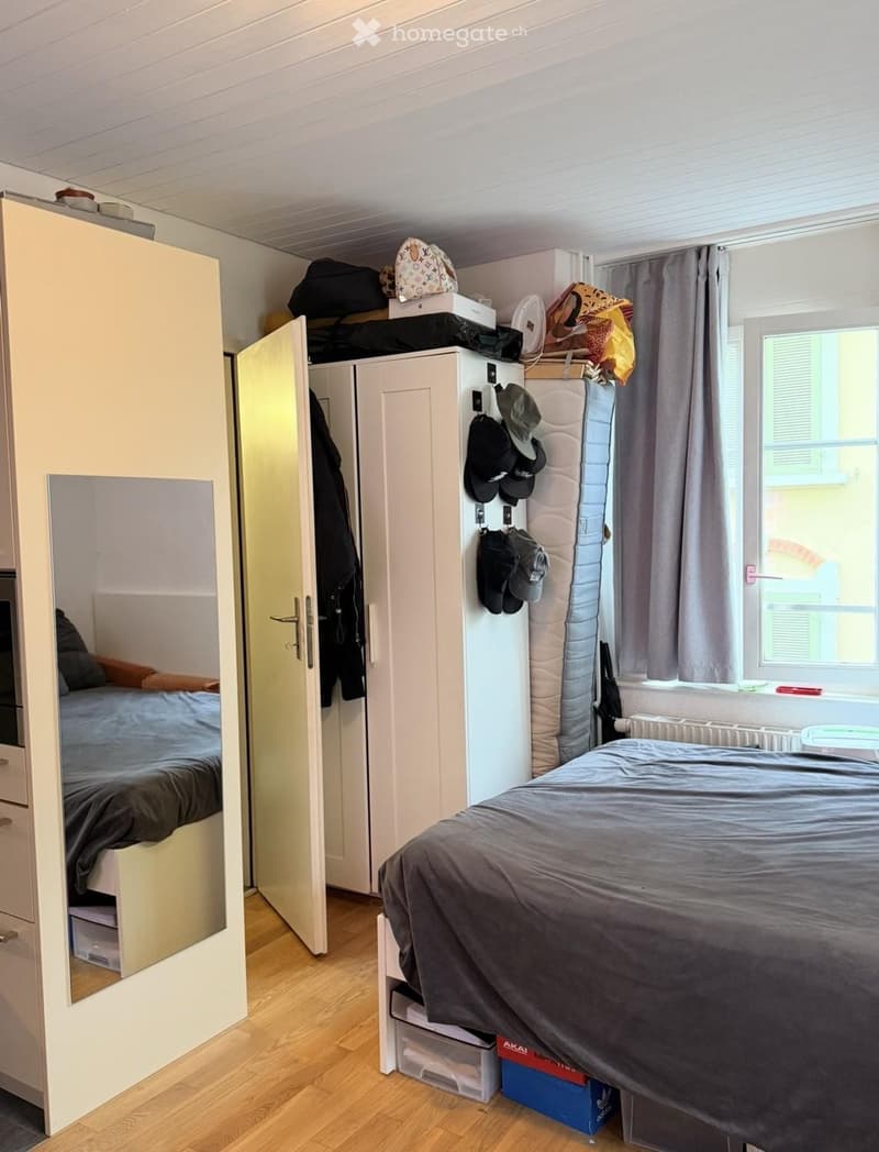 Cozy bedroom with two single beds, a wardrobe, a mirror, a hat rack, and storage boxes. The room features wooden flooring and a window with curtains.