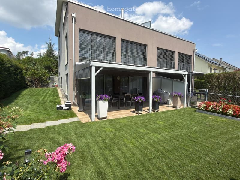 Well-maintained lawn with a modern house in the background, featuring a covered patio area with a dining set and flower pots.