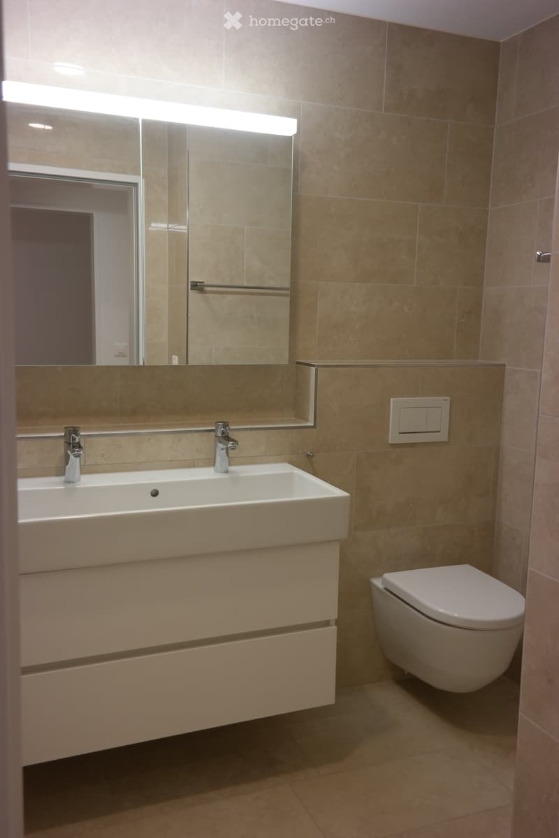 Modern bathroom with beige tiles, a large sink with dual faucets, a mirror with built-in lighting, and a toilet.