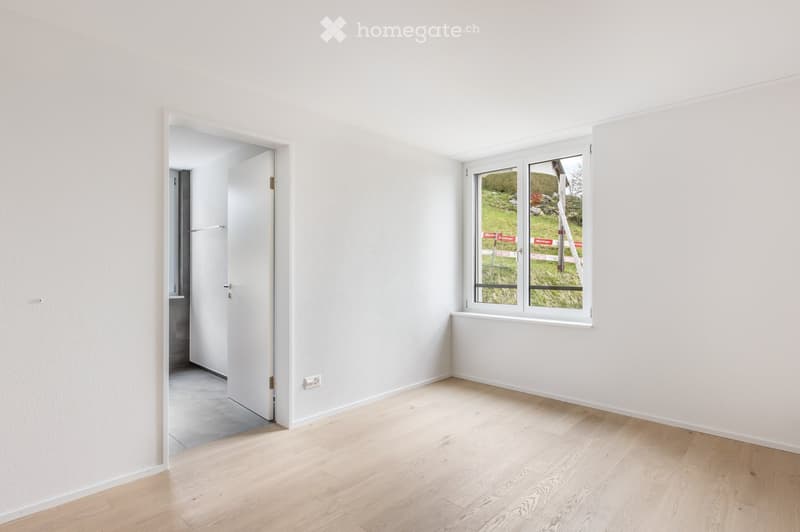 Empty room with light wooden flooring, white walls, and a large window showing a green outdoor view. A doorway leads to another room.