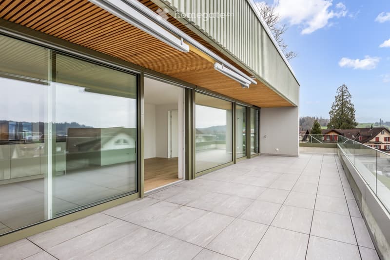 Modern balcony with large glass sliding doors, wooden ceiling, tiled floor, and a view of the surrounding landscape.