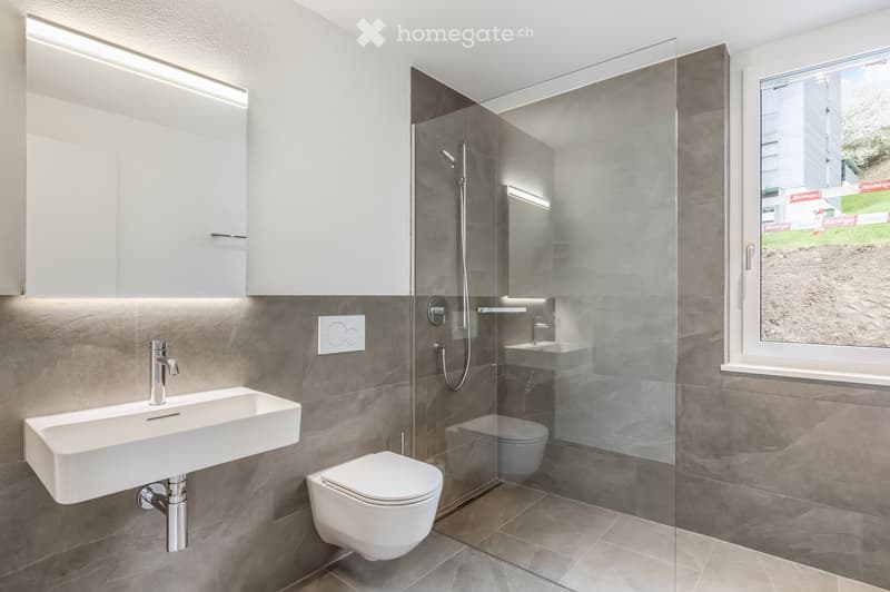 Modern bathroom with grey tiled walls, white sink, toilet, and glass-enclosed shower. Features built-in cabinets and a large window offering a view of the outside.