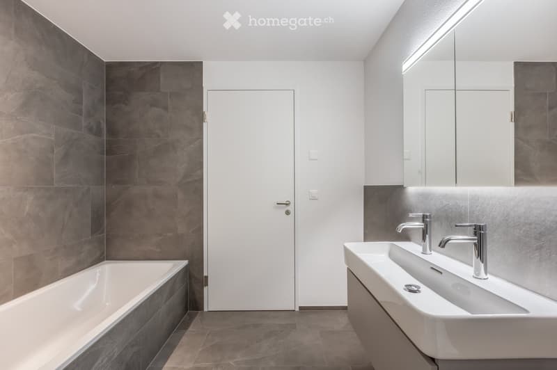 Modern bathroom with a white bathtub, double sink, and sleek chrome faucets. Dark grey tiles on walls and floor, white cabinetry, and a large mirror with integrated lighting.