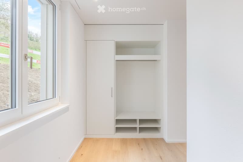Empty room with white walls, light wooden flooring, built-in closet with shelves, and a large window with a view of the outdoors.