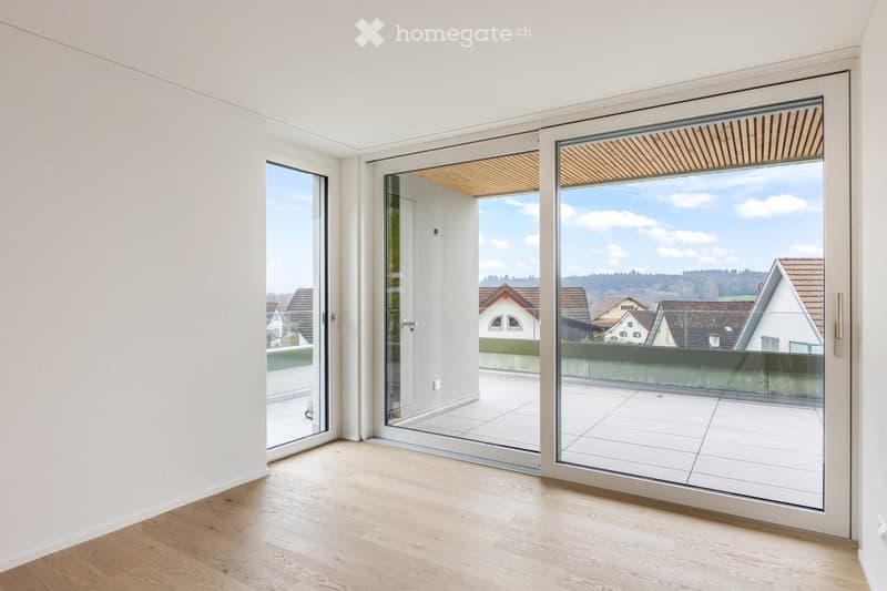 Bright, empty room with light wooden flooring and large sliding glass doors leading to a balcony with a view of the neighborhood.