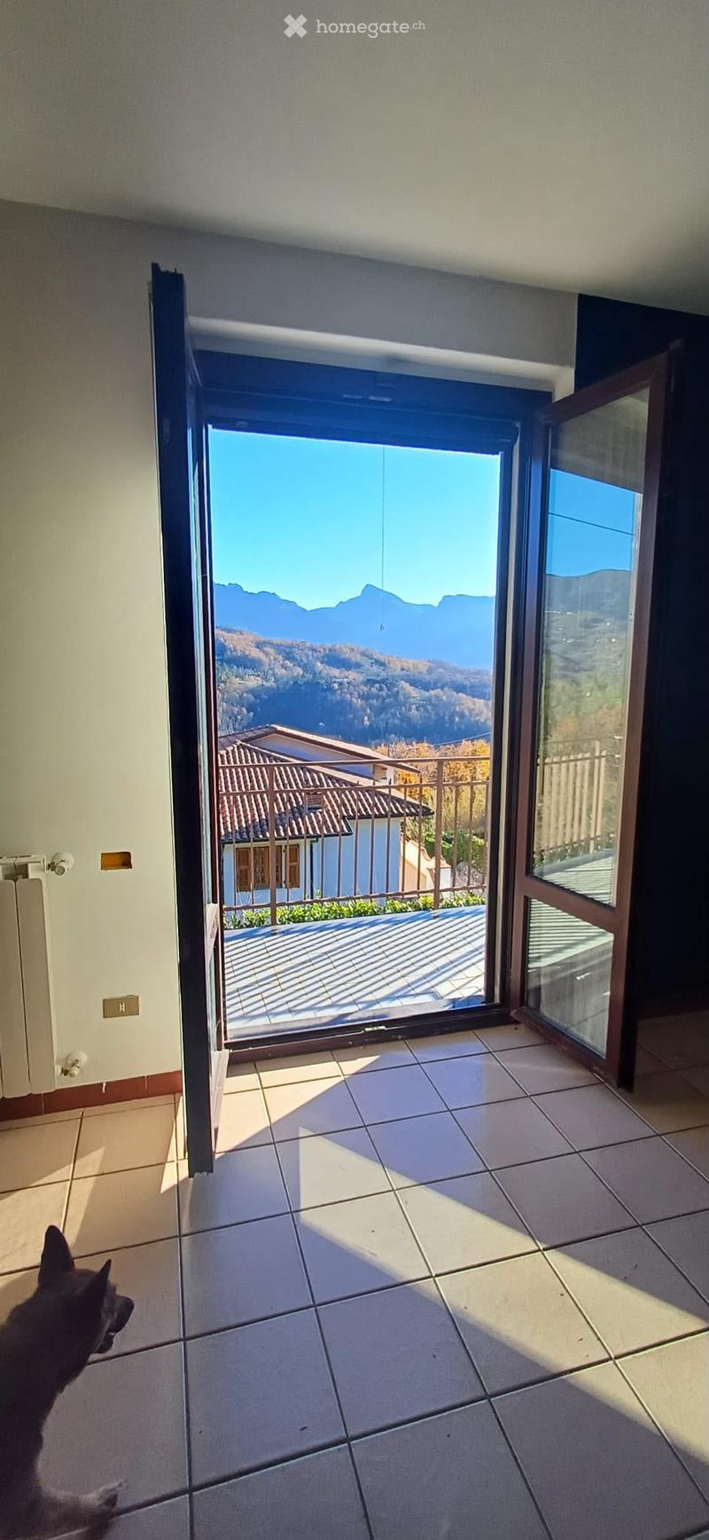 Open sliding glass door leading to a balcony with a scenic mountain view, tiled floor, and a dog visible in the foreground.