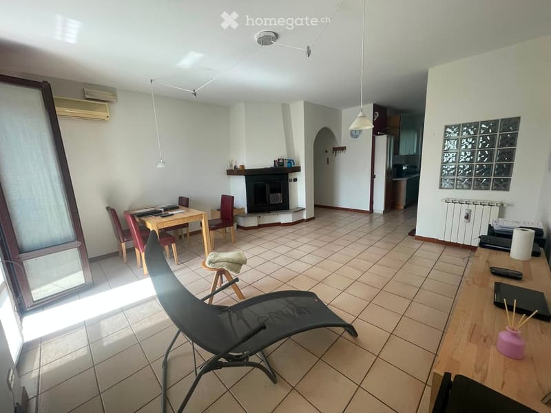 A well-lit living room with tiled flooring, a fireplace, a dining table with chairs, a lounge chair, and a desk with a laptop and accessories.