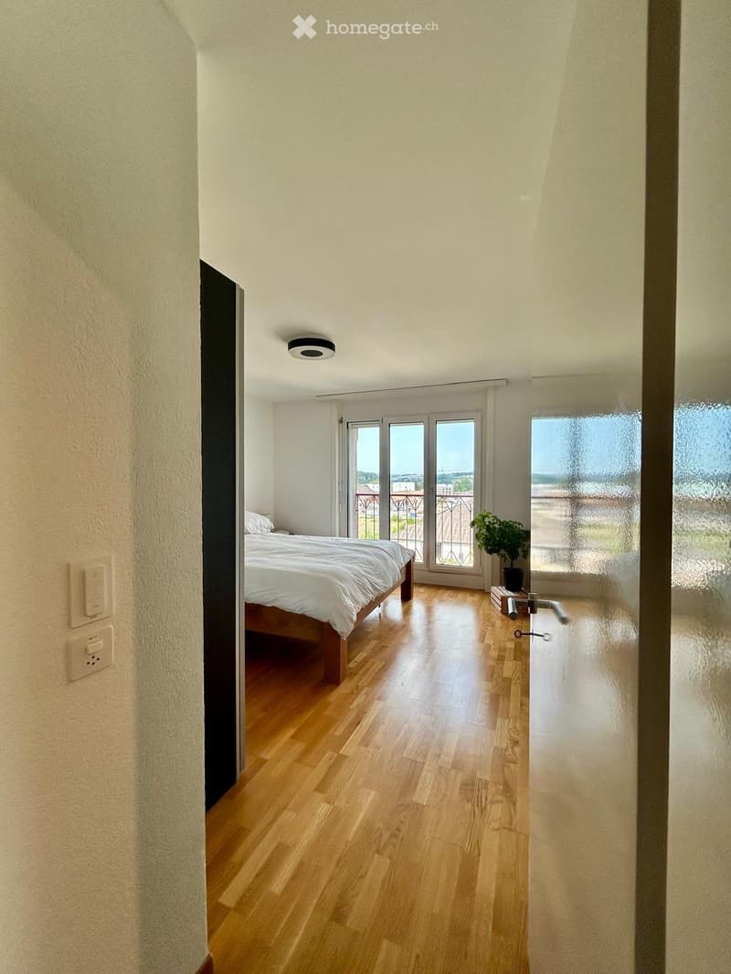 Bright bedroom with wooden flooring, large bed with white bedding, sliding glass doors leading to a balcony, and a small plant on the floor.