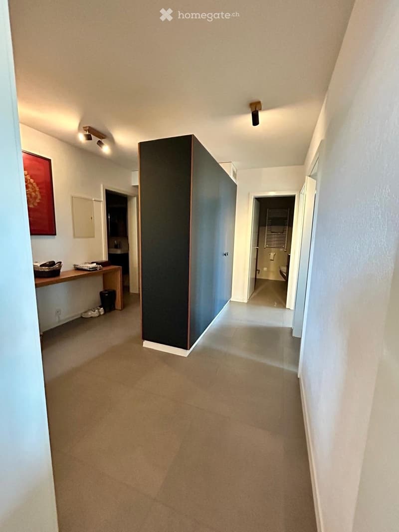 Carpeted hallway with white walls, recessed lighting, and a large dark partition in the center. Doors lead to adjacent rooms, including a bathroom and a small entryway area with a bench and shoes.