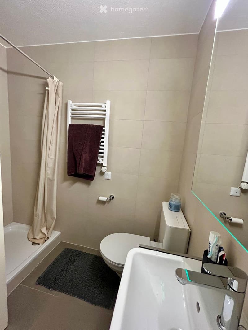 Modern bathroom with beige tiles, bathtub, toilet, sink, towel rack with a towel, and a small bath mat on the floor.