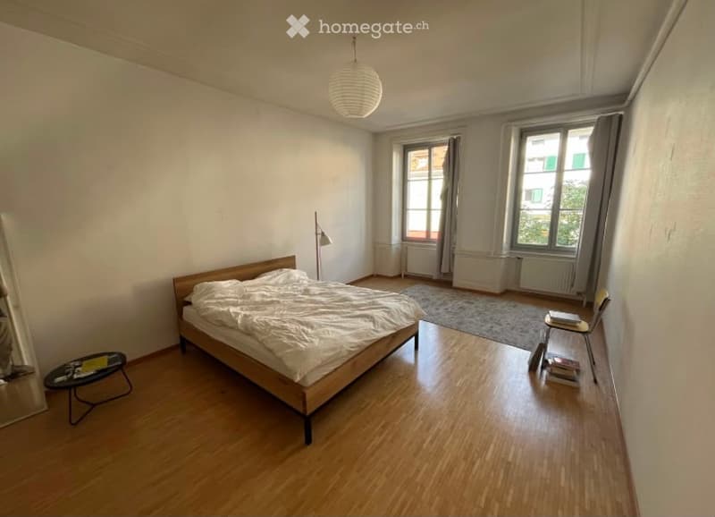 A modern, well-lit bedroom with wooden flooring, a large bed with white bedding, a small round table with books, a chair, and a floor lamp near the window.