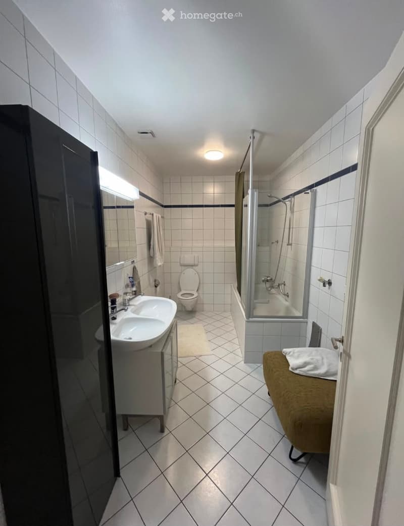 Modern bathroom with white tiled walls and floor, double sink, toilet, shower, and a small bench with a towel.