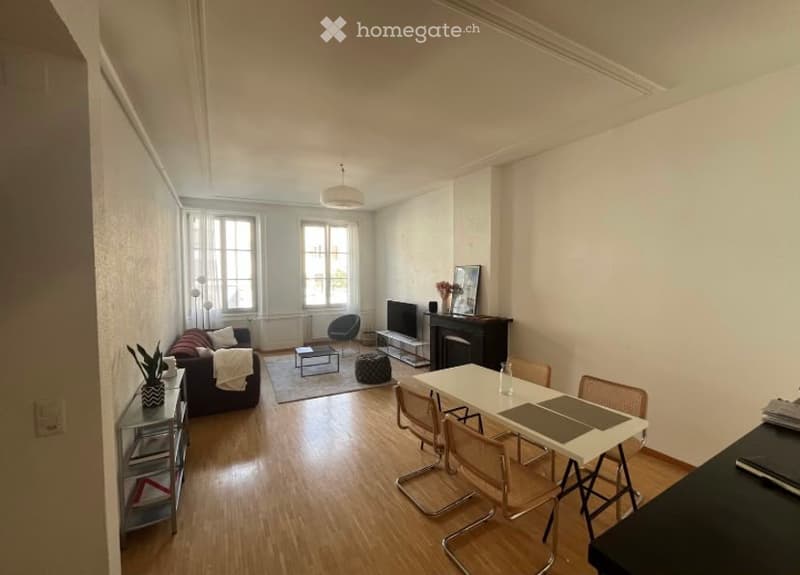 Bright living and dining area with wooden flooring, large windows, fireplace, sofa, coffee table, dining table with chairs, and decorative items.