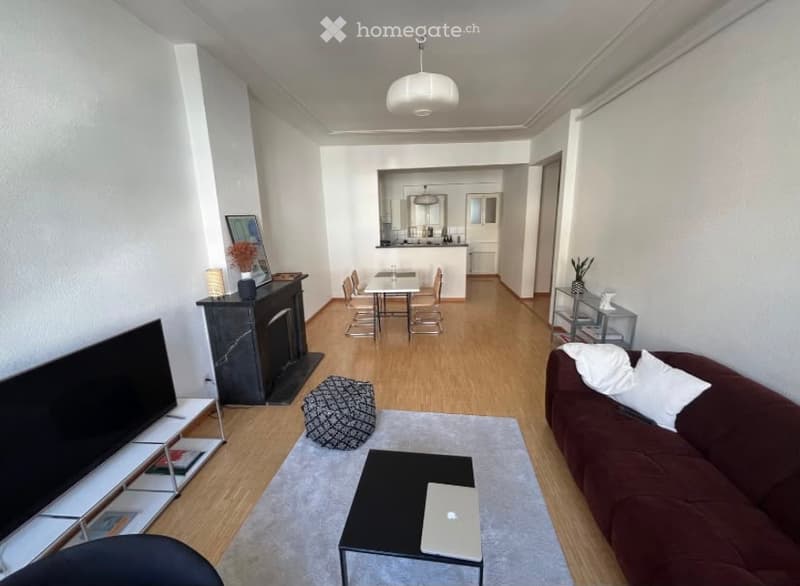 Modern living room with a brown sofa, coffee table, laptop, ottoman, fireplace, and dining area with chairs and a table. The room features light wooden flooring and white walls.