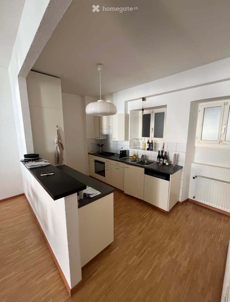 Modern kitchen with white cabinets, black countertops, wooden flooring, and a central island. Includes a sink, oven, and various kitchen items on the counter.