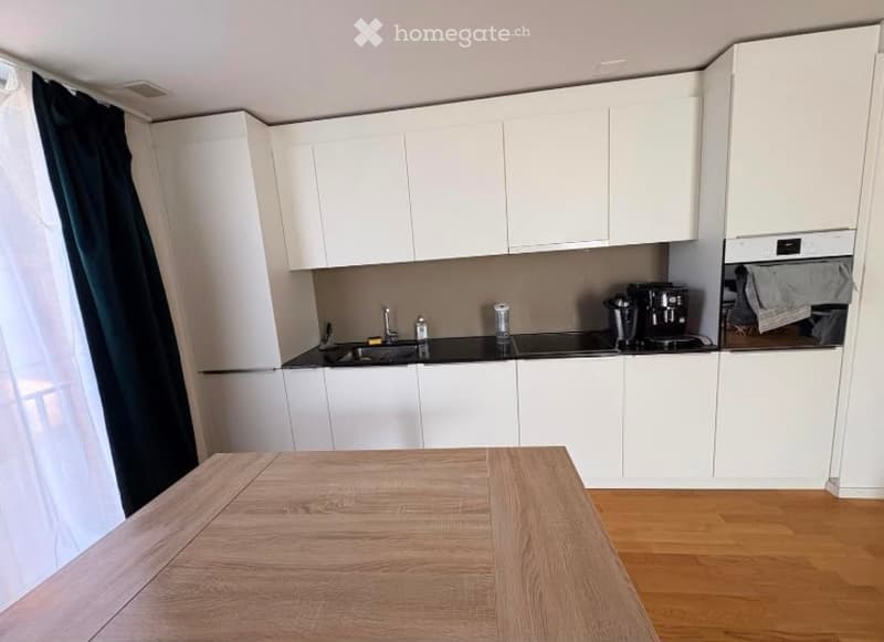 Modern kitchen with white cabinets, black countertops, and a wooden dining table. Appliances include a coffee maker, oven, and sink with a faucet.