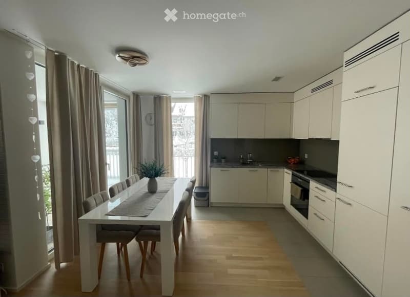 Open-plan dining and kitchen area with modern white cabinets, a dining table with chairs, and a small plant on the table. Large windows with curtains lead to a balcony.