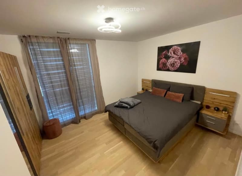 Modern bedroom with a large bed, nightstand, picture frame on the wall, and a small stool near the window. The room features wooden flooring and large windows with blinds.