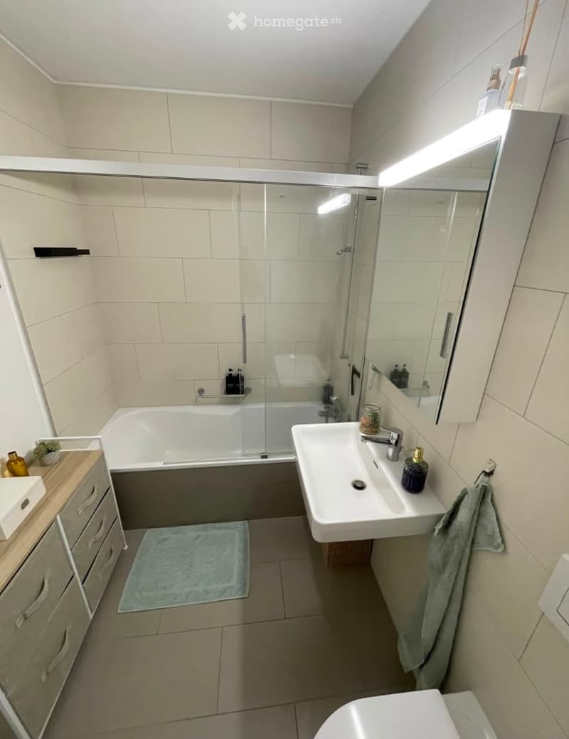 Modern bathroom with a bathtub, shower, sink, and toilet. Features a large mirror with built-in lighting, a cabinet with drawers, and a towel on the floor.