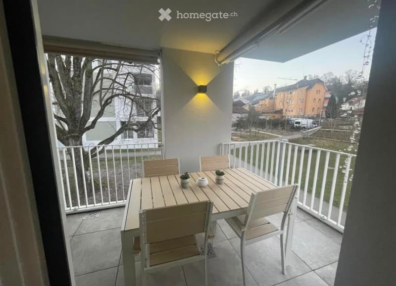 Balcony with a wooden dining table and chairs, small potted plants, white railing, and a view of the surrounding area.