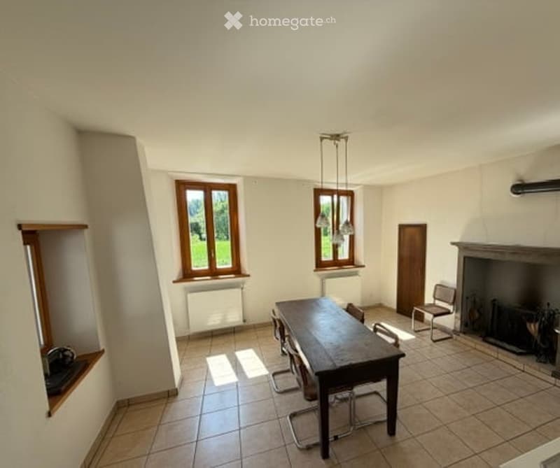 Bright dining area with wooden table, chairs, fireplace, and large windows overlooking a green landscape.