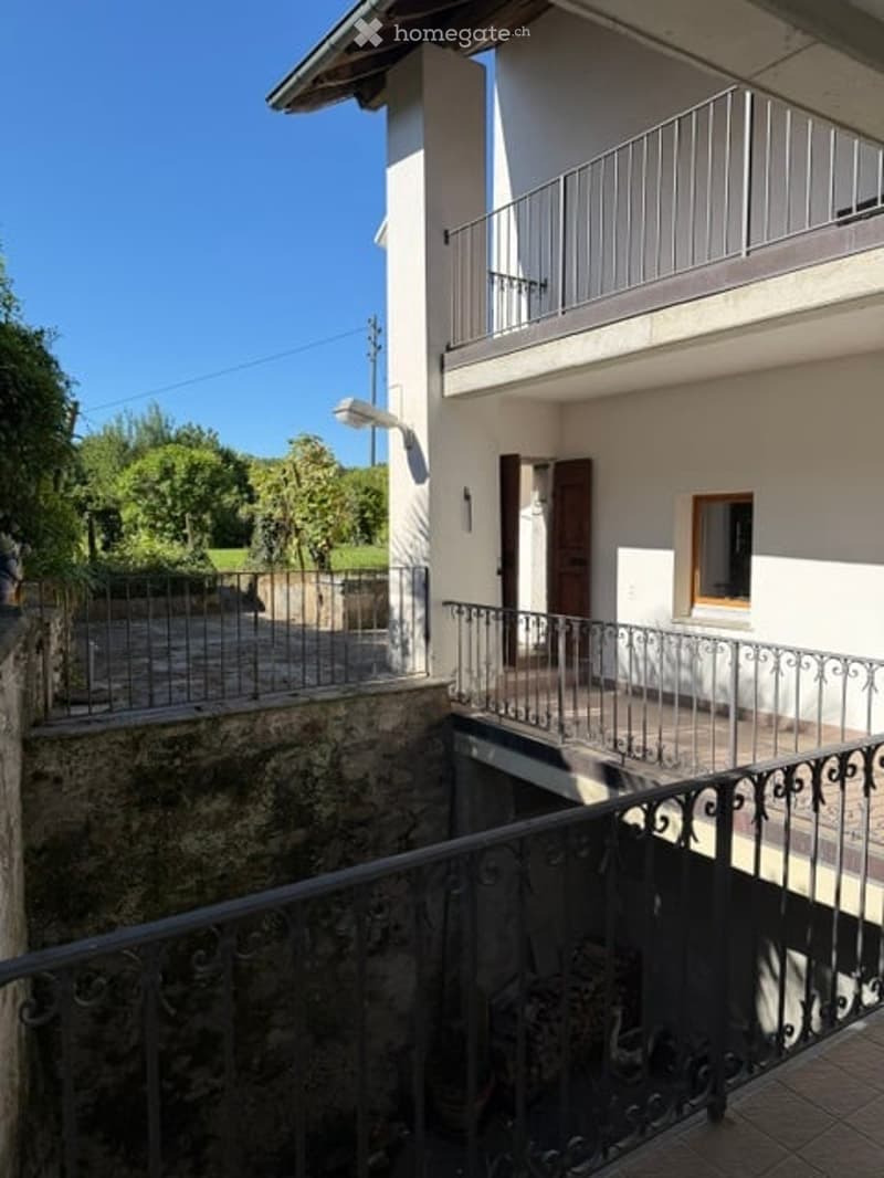Two-story residential building with a balcony, metal railings, and a stone lower level.