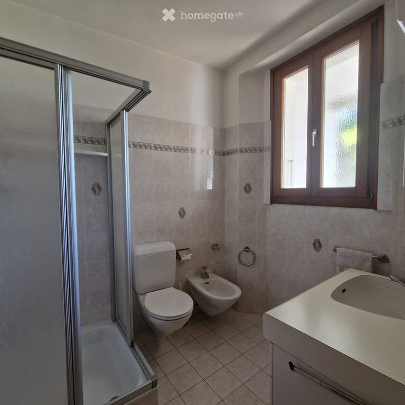 Modern bathroom with a toilet, bidet, sink, and a glass-enclosed shower. Light-colored tiles and a window provide natural light.
