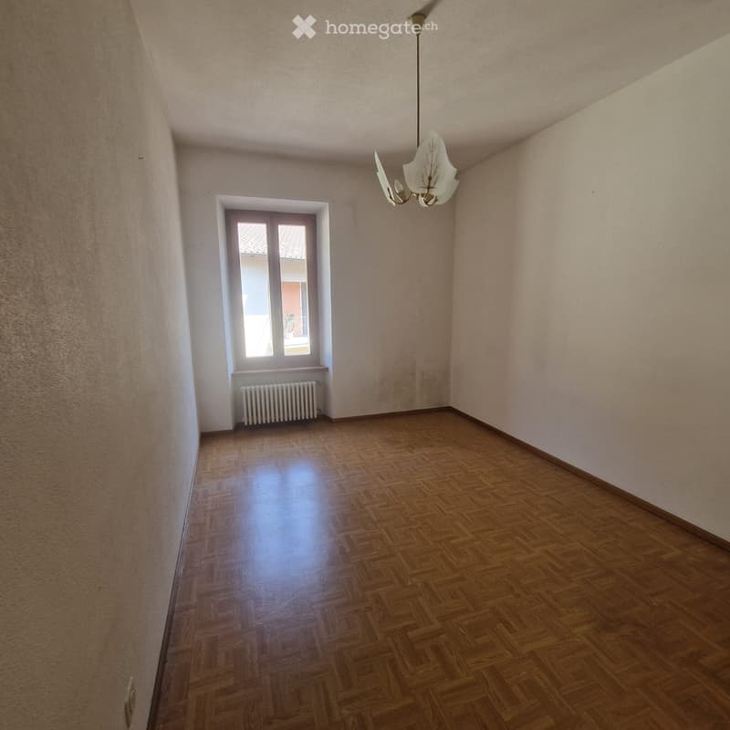 Empty room with wooden flooring, light-colored walls, and a single window with a radiator below it. A chandelier hangs from the ceiling.