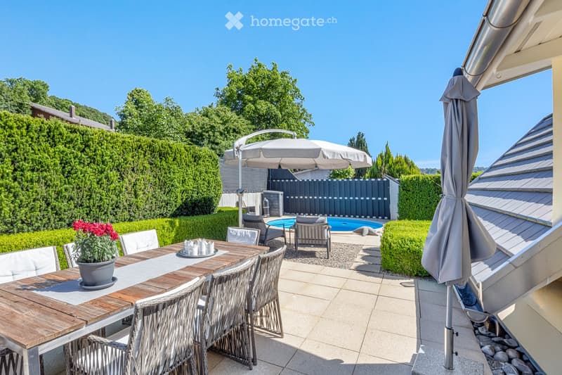 A well-maintained outdoor patio area with a wooden dining table, wicker chairs, a large umbrella, and a swimming pool in the background, surrounded by greenery and neatly trimmed hedges.