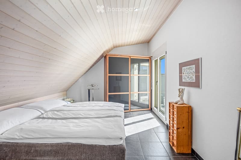 Modern bedroom with a slanted wooden ceiling, large bed with white bedding, nightstand, wardrobe with glass doors, and a decorative statue on a wooden chest of drawers.