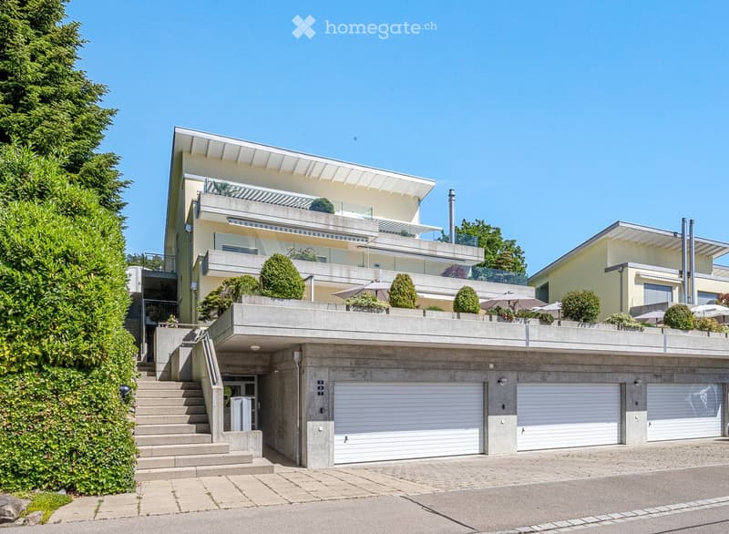 Modern two-story house with a raised ground floor, large garage, and balcony with greenery and outdoor seating.