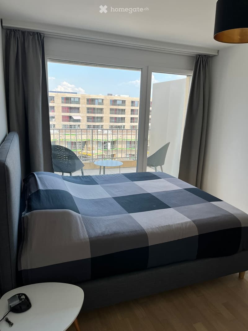 Modern bedroom with a large bed, checkered duvet, two chairs, small table, and large windows with a view of a building and balcony.